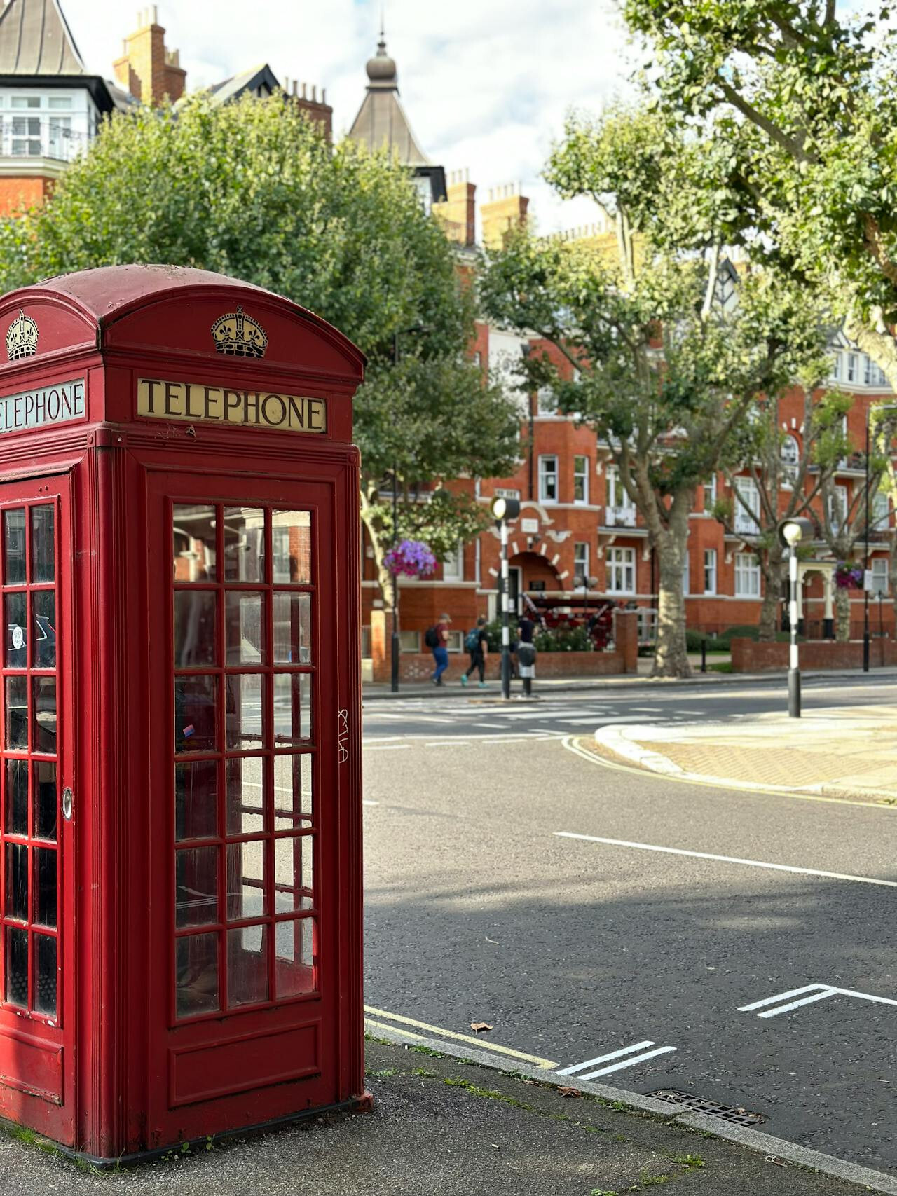 Κλασική κόκκινη τηλεφωνική καμπίνα σε γωνία δρόμου στο Λονδίνο, με φόντο βικτοριανά κτίρια και καταπράσινα δέντρα σε ήρεμη, ηλιόλουστη μέρα.