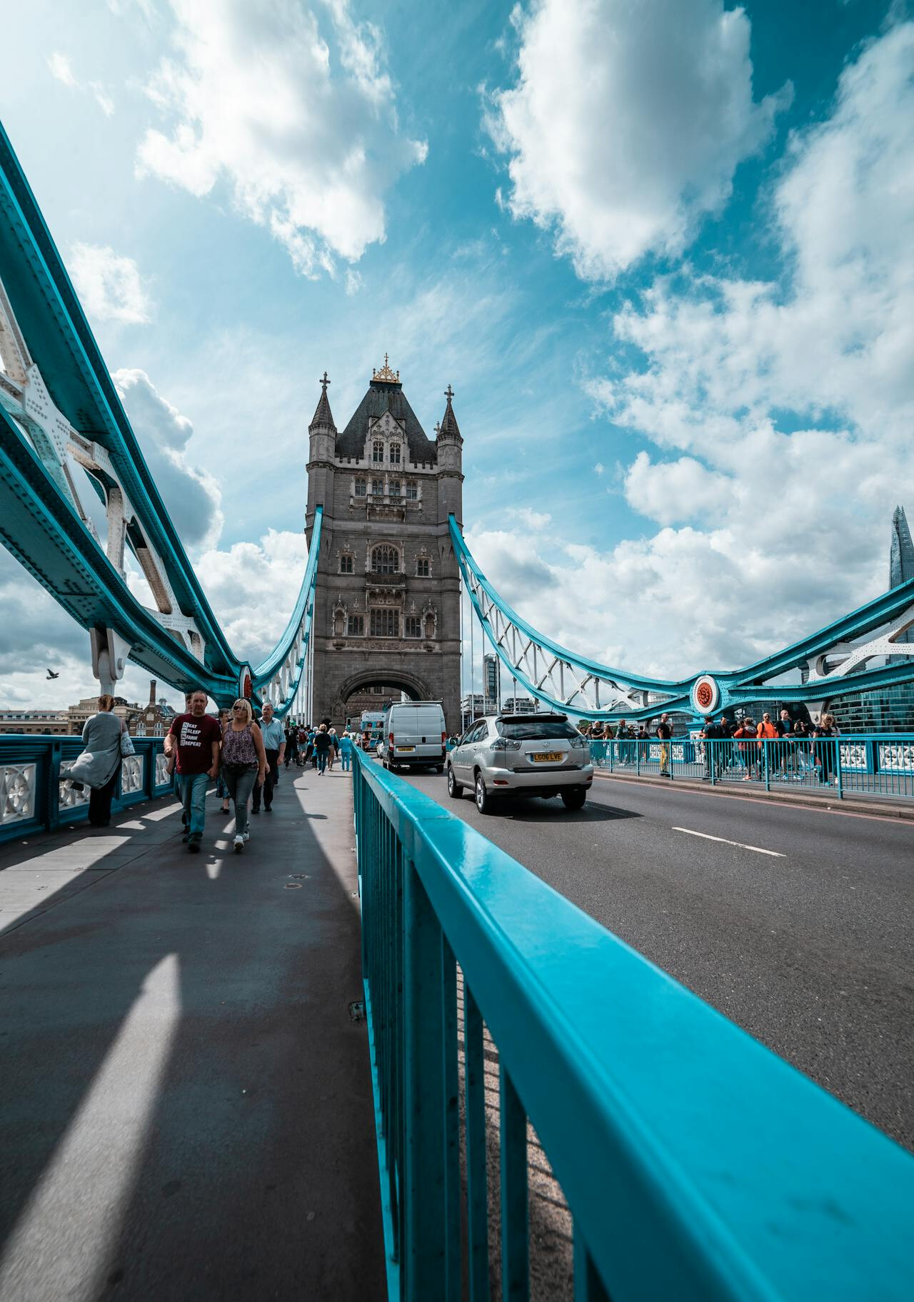 Πεζοί και αυτοκίνητα διασχίζουν την iconic Tower Bridge του Λονδίνου κάτω από έναν θεαματικά συννεφιασμένο ουρανό, με το μπλε στηθαίο να οδηγεί το βλέμμα στον μεσαιωνικό πύργο της γέφυρας.